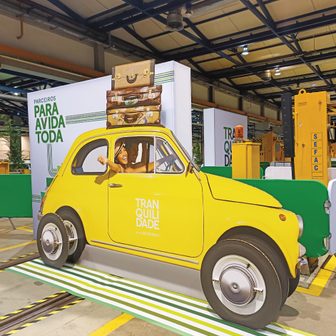 Carro amarelo decorativo com malas empilhadas no topo, com destaque para a palavra 'Tranquilidade', em um evento de design corporativo.