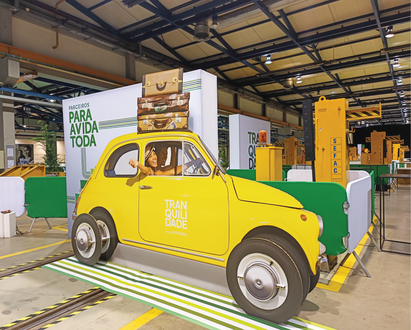 Carro amarelo decorativo com malas empilhadas no topo, com destaque para a palavra 'Tranquilidade', em um evento de design corporativo.